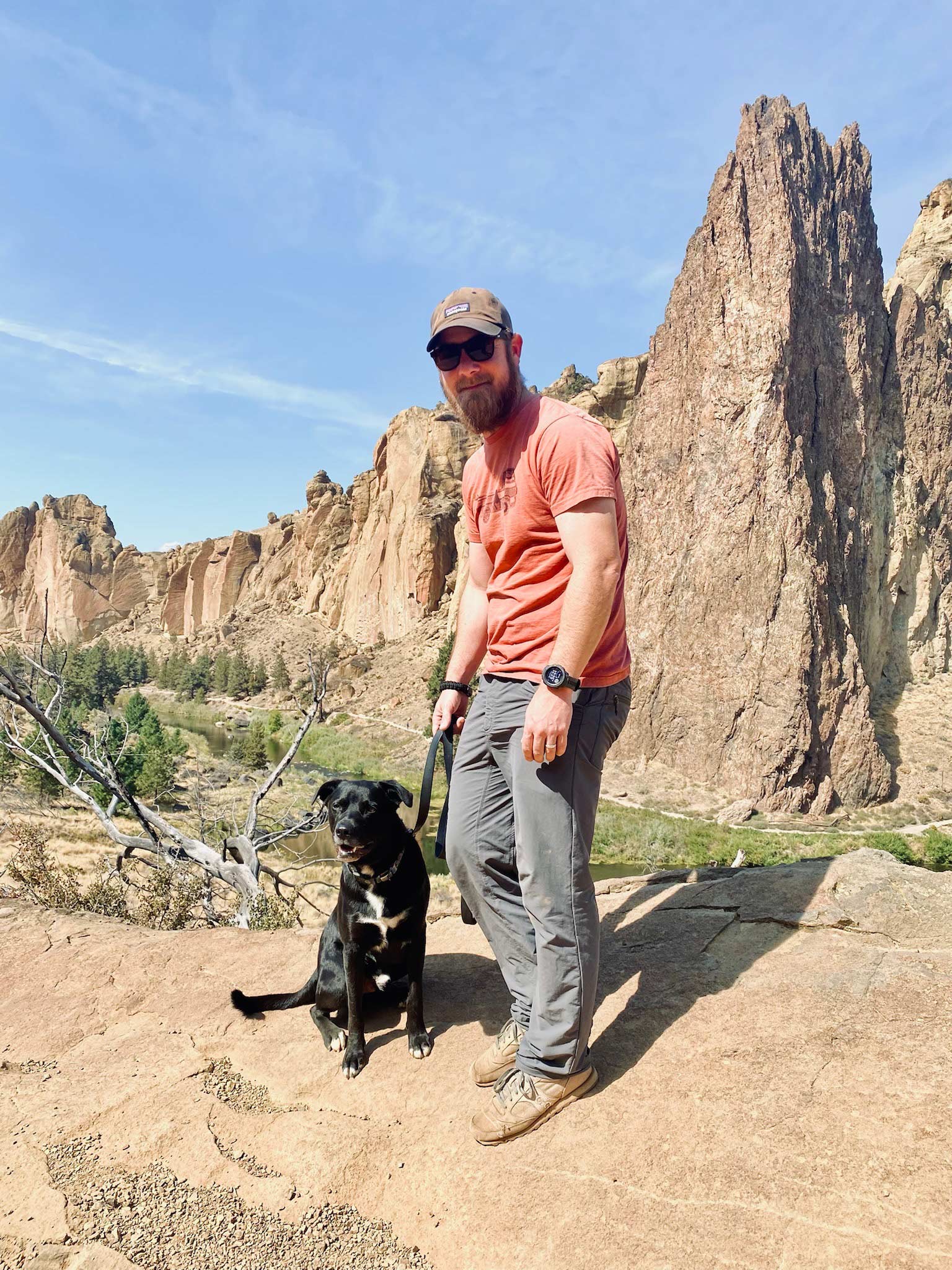 A man in an orange shirt, sunglasses, gray pants, and a watch holding a dog in front of a rock spire in the background