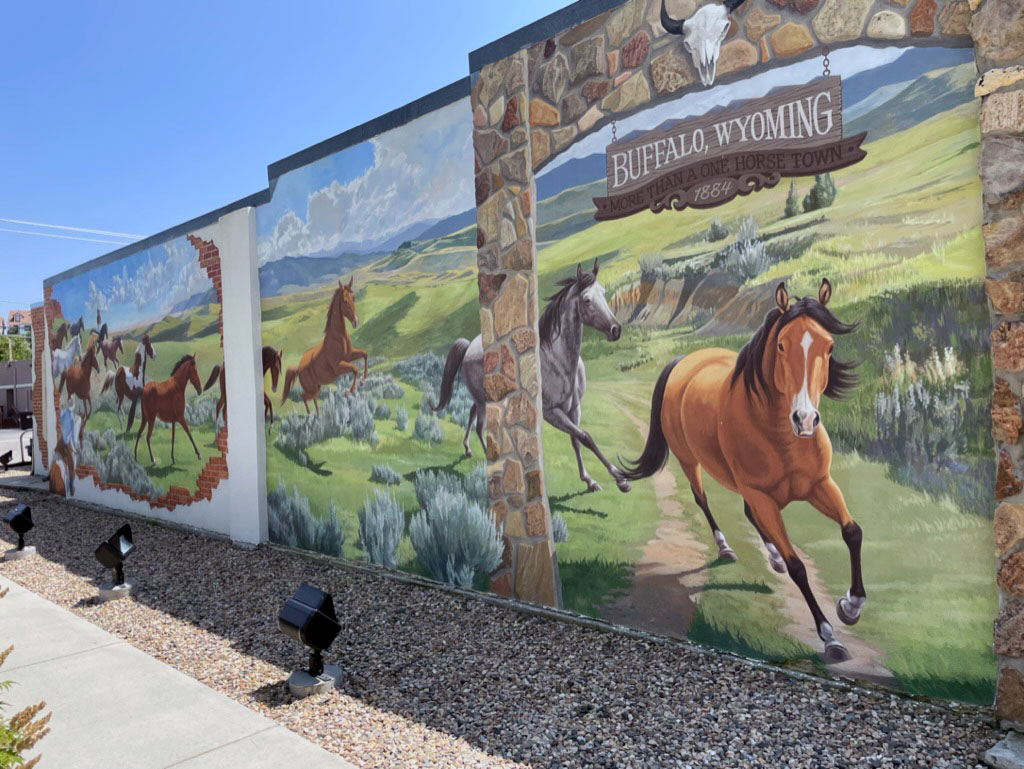 A mural on the side of a building showing horses galloping among green pastures and sage