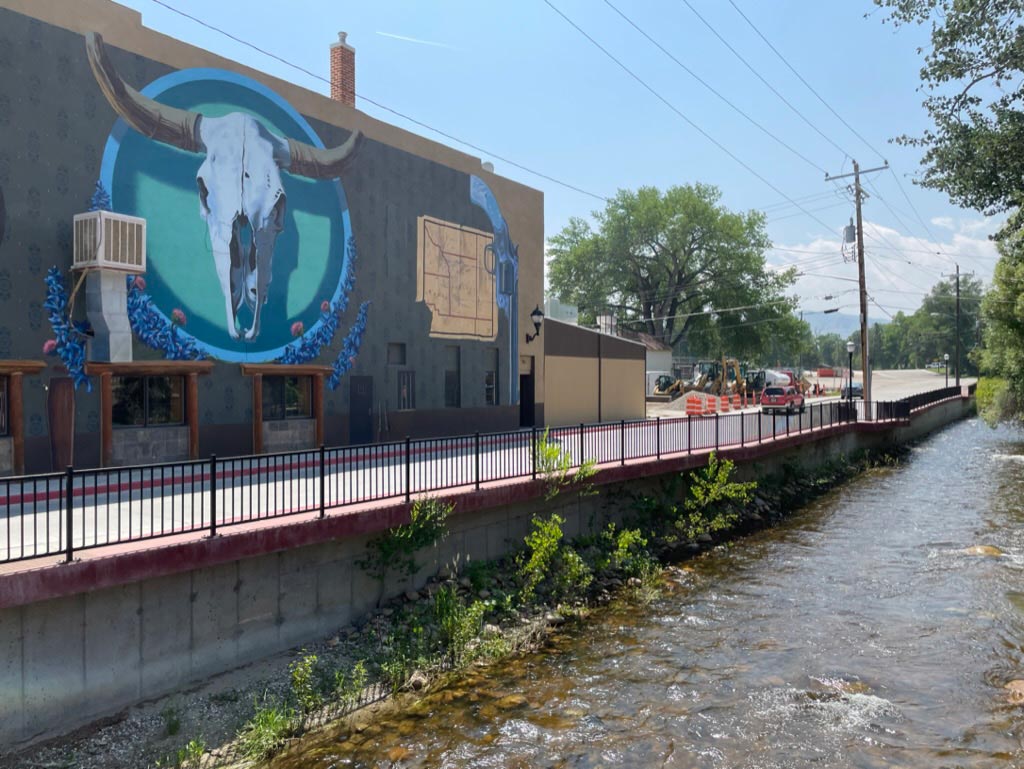 A mural of a buffalo skull on the side of a building aligned along a path and creek